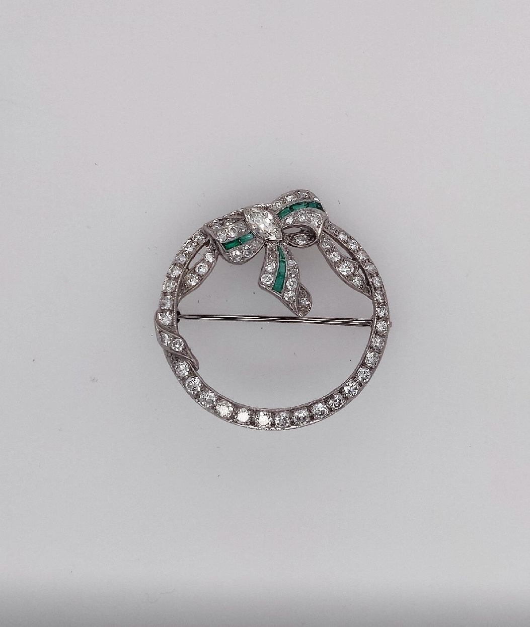 Platinum Art Deco Ribbon and Bow Circle Brooch with Diamonds and Emeralds
Brooch Contains one Marquise Shaped Diamond weighing approximately 0.20CT J/VS1
58 early full cut damonds; approximately 1.8CTTW G-H/VS
8 Calibre Cut Emeralds